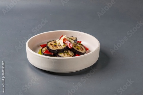 Kyopolou in white ceramic bowl filled with sliced eggplant, peppers, olives, topped with red onions, herbs. Concept of roasted eggplant pepper spread with garlic, vibrant dish.
