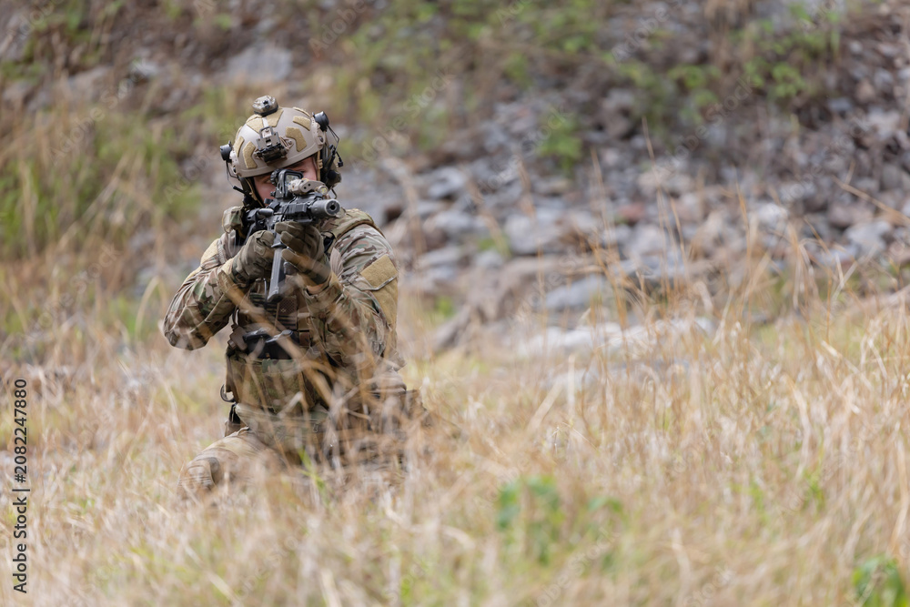Fototapeta premium Soldier in military gear aims weapon while crouching in tall grass