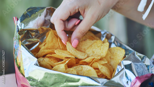 Woman's hand with pink nail polish picking a potato chip from an opened foil bag of chips