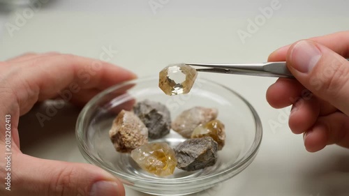 Hand with tweezers carefully selecting small amber gems from clear dish with shallow depth of field
