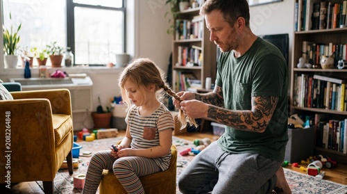 Affectionate tattooed father braiding his young daughter's hair while sitting on the living room couch.