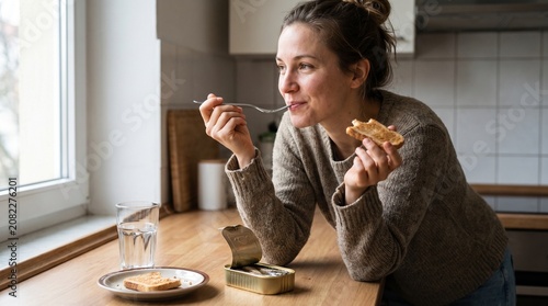 Young Caucasian woman happily eating a quick sardine meal against a bright kitchen window background