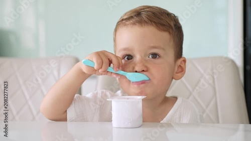 Little boy with light brown hair holds a blue spoon to his mouth while eating yogurt from a small white container, seated at a white table in a well-lit kitchen. Healthy nutrition concept. Footage