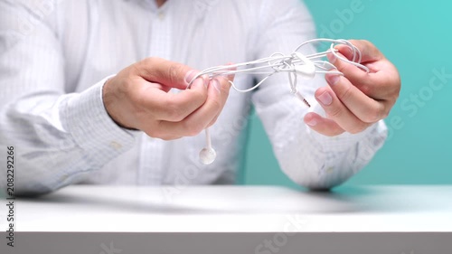 Person's hands carefully manipulating a knot of white tangled earphones, showing frustration with a common daily problem against a blue background and white table