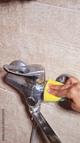 Hand cleaning a bathroom faucet or water tap with a yellow sponge, removing limescale and water stains from chrome tap, household cleaning and hygiene concept