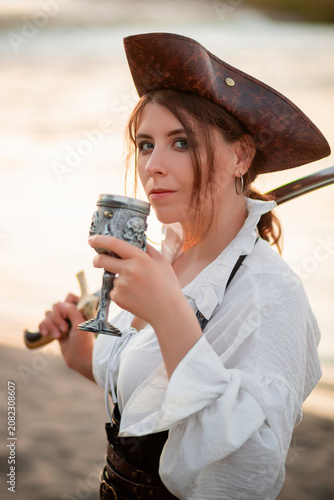 Foto Portrait of a young attractive woman in a pirate costume posing with a saber and a glass in her hands against the background of the shore and water