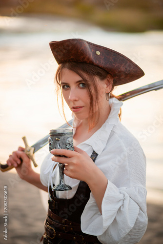 Schilderij op canvas Portrait of a young attractive woman in a pirate costume posing with a saber and a glass in her hands against the background of the shore and water