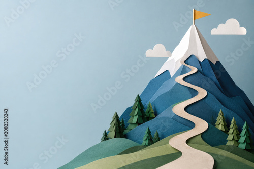 Paper mountain with winding path and summit flag. Space for text.