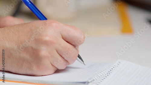 Close-up of a man's hand writing with a blue pen in a spiral notebook. Male hand taking notes or signing a document on lined paper, business and education concept