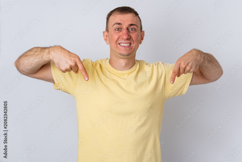 Fototapeta premium Cheerful adult man standing proudly in a yellow t shirt, smiling while pointing both index fingers down, indicating a space for important advertising content