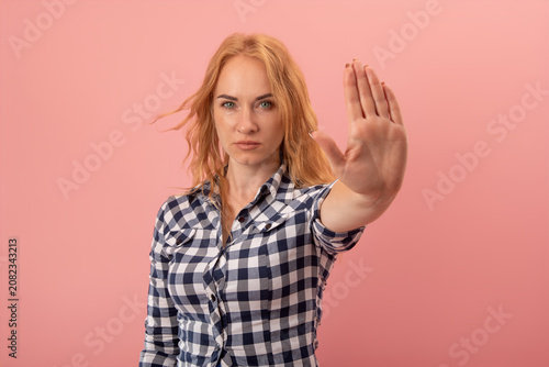 Young woman with blonde hair wearing a plaid shirt making a stop gesture with her open palm, signaling refusal, boundary setting, or warning on a pink background