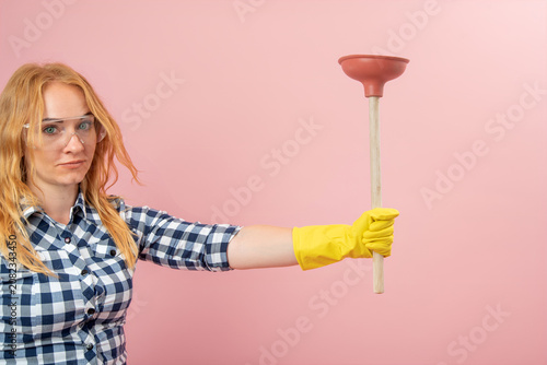 Young woman wearing a plaid shirt and protective eyewear with a serious expression, holding a toilet plunger, representing concepts of plumbing, cleaning, and DIY home repair