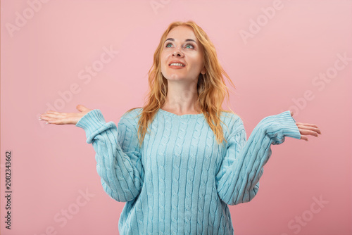 Smiling woman with reddish hair and blue eyes looking upwards, holding both hands open, palms facing up, against a pink background