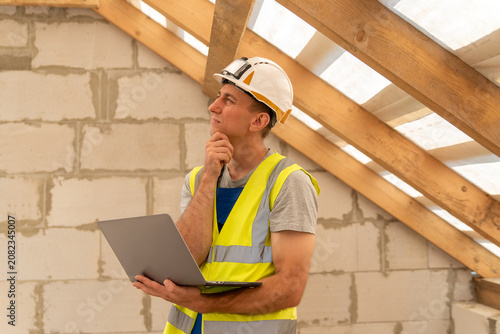 Construction worker wearing a hard hat and safety vest, holding a laptop and contemplating plans in an unfinished building with wooden roof beams and brick walls