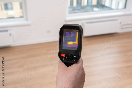 Hand holds a thermal camera. Screen shows color-coded heat patterns. Room has wooden flooring and windows. Professionals use it for energy audits. It highlights thermal imaging applications