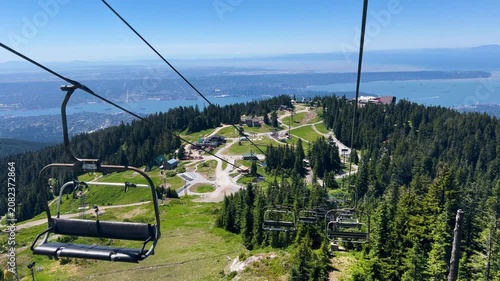 A chairlift descends over the Grouse Mountain summit toward its lodge and ski runs, Vancouver, Burrard Inlet and the ocean spread below on a clear summer day