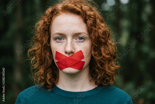 Close-up portrait of a young woman with red tape over her mouth, symbolizing censorship, silenced voices, freedom of speech, human rights, oppression, and social justice.