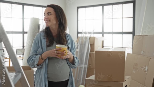 Woman holding a yellow coffee mug amid stacked moving boxes and ladder in a sunlit building room, smiling and cradling her bump; hopeful anticipation.
