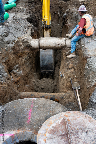 Excavator digging a deep narrow ditch in the dirt to work on underground utilities, construction worker in trench guiding machine operator around old concrete pipe
