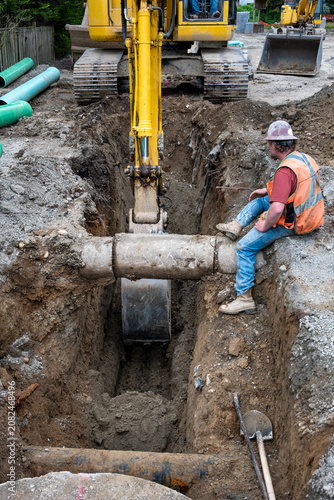 Excavator digging a deep narrow ditch in the dirt to work on underground utilities, construction worker in trench guiding machine operator around old concrete pipe
