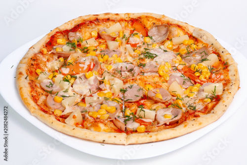 Whole baked pizza with sausages corn, tomatoes pineapple and herbs served on a white plate.