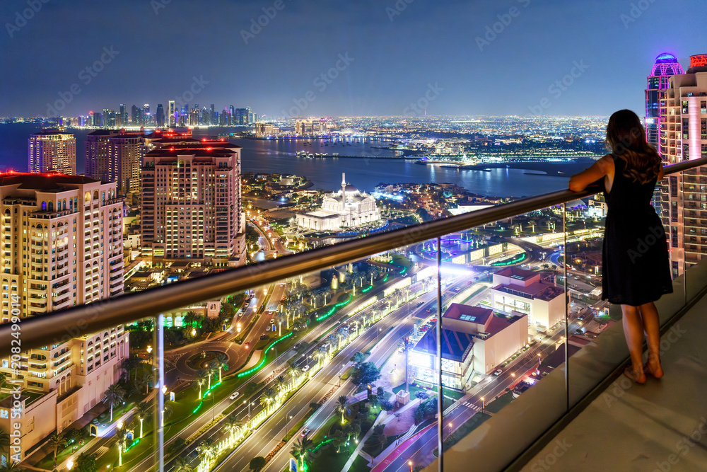 Fototapeta premium A young woman stands on a high rise terrace at night overlooking the illuminated Doha Pearl, downtown skyline, and the waters of the Persian Gulf in Doha Qatar.