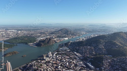 Aerial panoramic view of Vitória, Espírito Santo, Brazil, featuring Vitória Bay, islands, mountains, urban skyline, coastline, and clear blue sky captured by drone.
