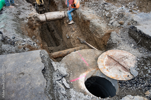 Underground utilities construction project, precast concrete stormwater system access vault in dirt trench under road, shovel and tamper bar, workman in trench guiding excavator around concrete pipe