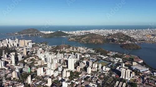 Aerial panoramic view of Vitória, Espírito Santo, Brazil, featuring Vitória Bay, islands, mountains, urban skyline, coastline, and clear blue sky captured by drone.