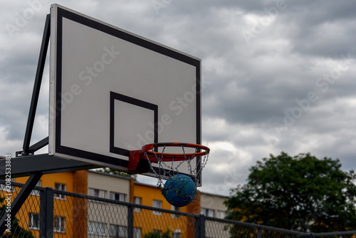Basketball falling through a metal chain net of an outdoor hoop on a streetball court against a dramatic cloudy sky.