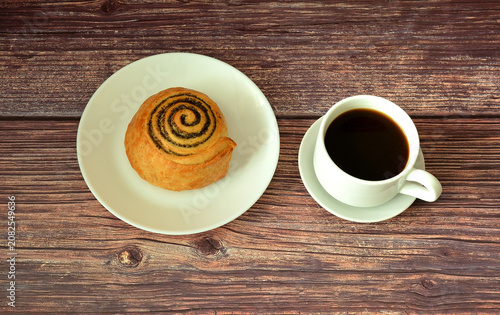 Cup of hot black coffee on a saucer and a plate with a freshly twisted poppy seed bun made of puff pastry on a wooden table.