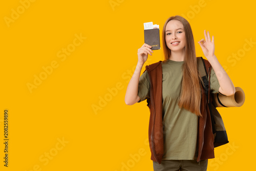 Smiling young woman with passport and ticket showing OK gesture on yellow bac...