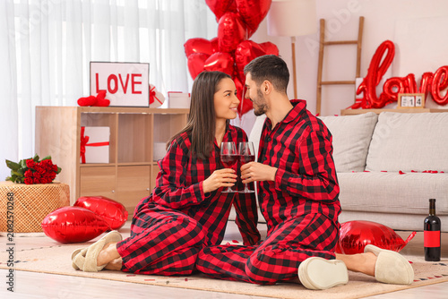 Young couple in checkered pajamas drinking wine at home. Valentines Day celeb...
