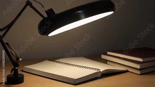 open notebook and stack of books under desk lamp on wooden table