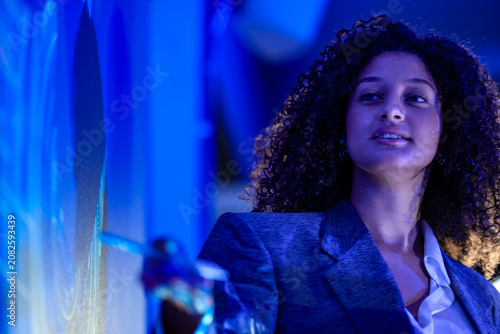 Businesswoman with curly hair outlines the roadmap with open hands rim lit in gold while speaking beside a deep blue wall inside a futuristic office illuminated by glowing blue projections