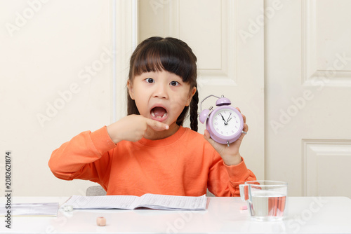 Obraz na plátně A surprised Asian little girl points to the alarm clock, seemingly expressing the concept of being late or time pressure