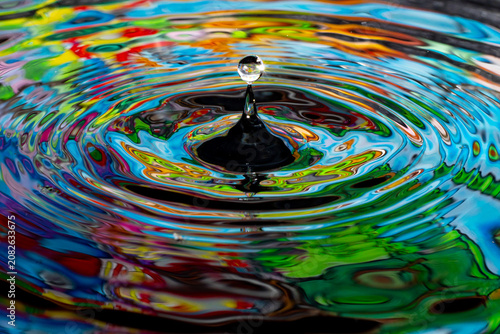 Small water droplets bounce off the surface of the liquid, creating ripples that emit abstract colorful reflections.