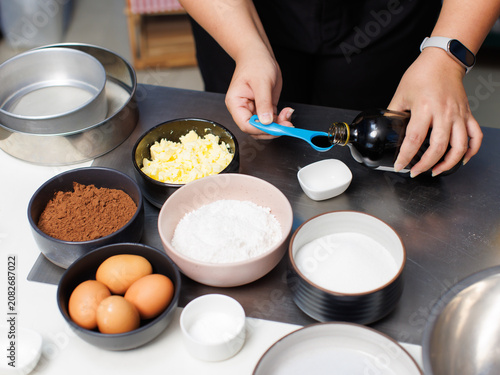 Asian woman pouring vanilla measuring spoon baking ingredients prepared dessert making cocoa powder sugar butter egg salt in bowls professional bakery fresh ready mixing homemade cake chocolate recipe