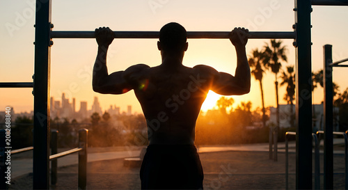 Silhouette of a muscular man performing a pull-up against a vibrant sunset over a city skyline.