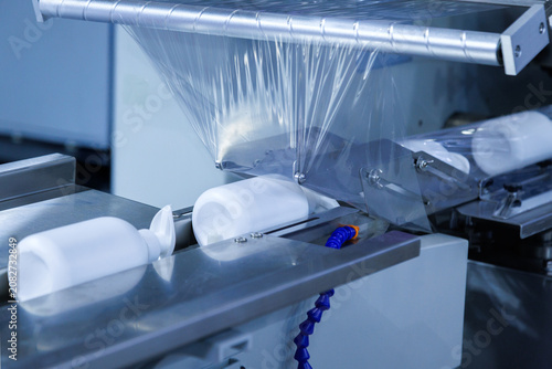 automatic shrink wrapping machine applying clear plastic film to white lotion cream pump bottles on conveyor belt in cosmetic factory. Industrial packaging, processing technology, and manufacturing.