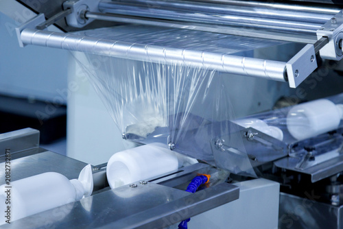 automatic shrink wrapping machine applying clear plastic film to white lotion cream pump bottles on conveyor belt in cosmetic factory. Industrial packaging, processing technology, and manufacturing.