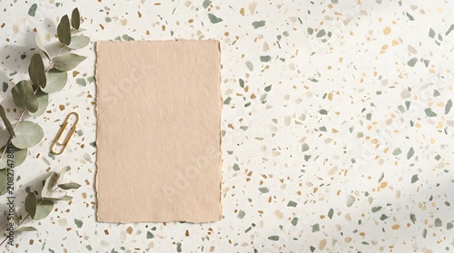 Blank deckle edge paper card with eucalyptus branch and gold paperclip on a modern terrazzo stone background flat lay
