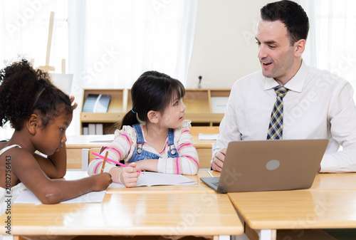 Fotografie Teacher talks with schoolgirl beside laptop while classmate studies nearby, showing communication, guidance and active learning during classroom lesson in elementary education with support