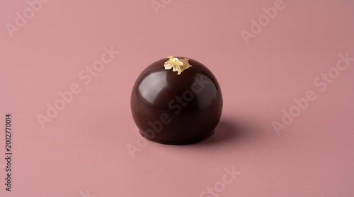 A glossy, dark chocolate dome mousse dessert topped with elegant edible gold leaf, centered against a minimalist dusty pink background.