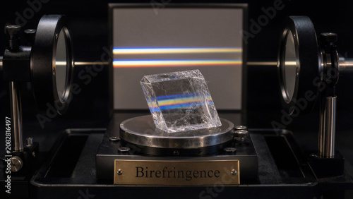Study shows birefringence in crystal using polarized light with setup featuring lenses and a transparent cube on a rotating stage