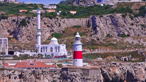 Drone footage panning telephoto across Europa Point — Ibrahim-al-Ibrahim Mosque's white dome and minaret beside the red and white lighthouse, limestone cliff behind.