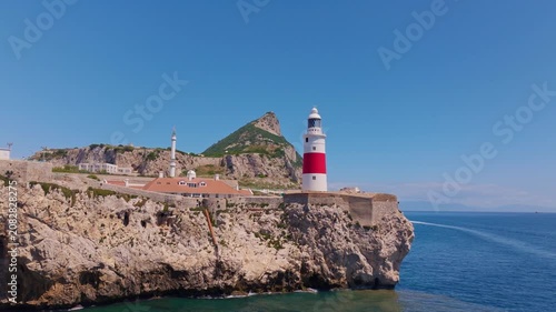 Drone footage approaching and ascending past Europa Point lighthouse — red and white tower, Ibrahim-al-Ibrahim Mosque, limestone cliffs, and Mediterranean Strait beyond.