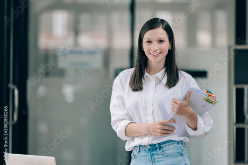 Young woman in casual outfit holding clipboard standing in office smiling