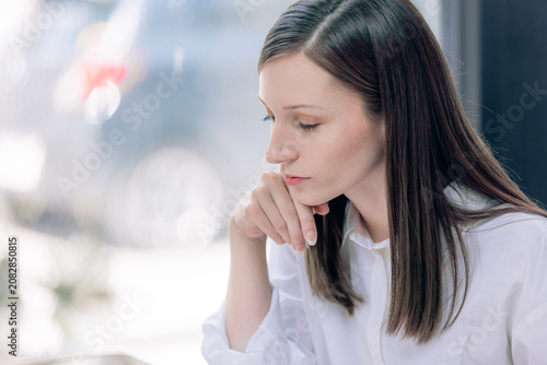 Thoughtful woman with long hair sitting indoors looking down in white shirt