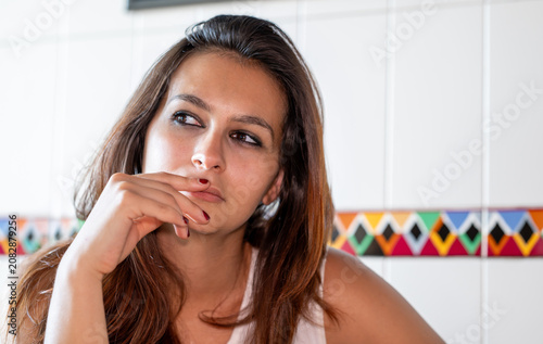 Young woman with brown hair and a serious expression deeply thinking, resting hand on her chin, looking away from the camera, against a white tiled wall
